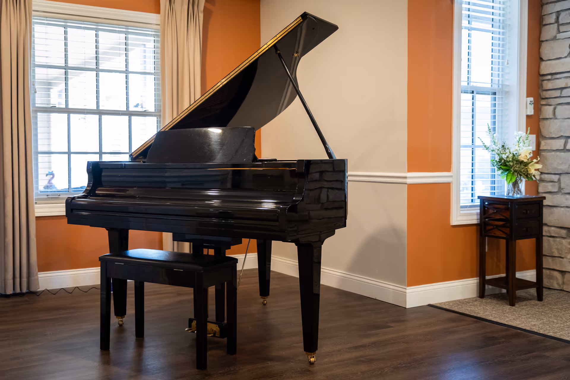 A black grand piano with a bench in a warmly decorated common room featuring windows, orange walls, wood floors, and a small table with flowers.