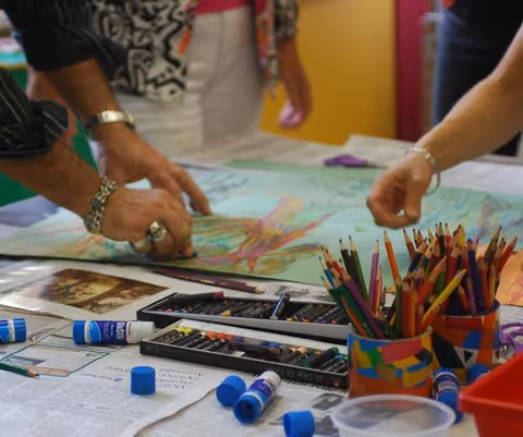 Two people working on a colorful drawing or painting on a table covered with newspaper, surrounded by colored pencils, oil pastels, and glue sticks.