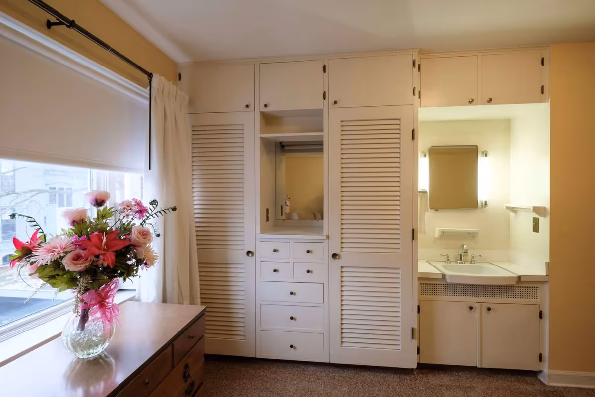 Interior view of a room with a large window on the left covered by a white roller shade and curtain. A wooden dresser with a glass vase holding a bouquet of pink and red flowers is in front of the window. On the right side, there is a built-in white cabinet with louvered doors and drawers, and an adjacent small sink area with a mirror and lights on the wall.