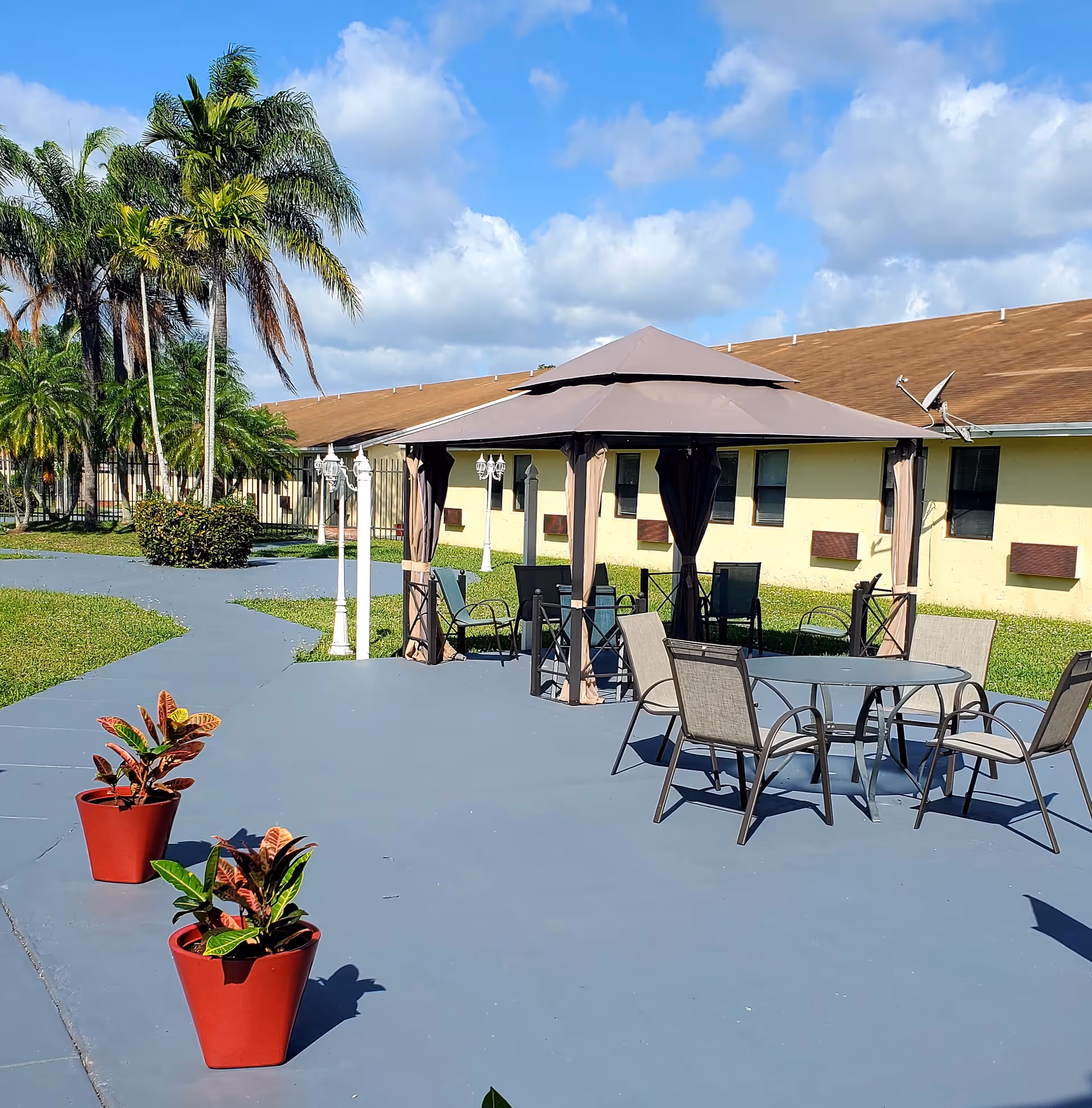 Outdoor patio area at Sunny Hills Assisted Living Facility Homestead with a gazebo, several chairs, a round glass table, two red potted plants, palm trees, and a yellow building with brown roof in the background under a partly cloudy blue sky.