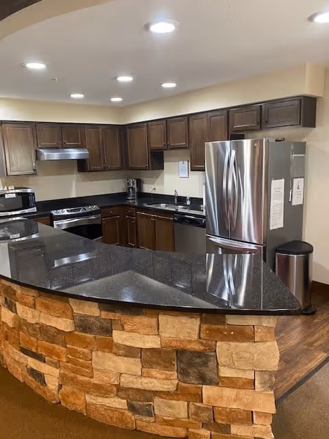 Modern kitchen with dark wooden cabinets, stainless steel refrigerator, stove, microwave, and dishwasher. The kitchen features a black granite countertop with a stone brick base and recessed ceiling lights.