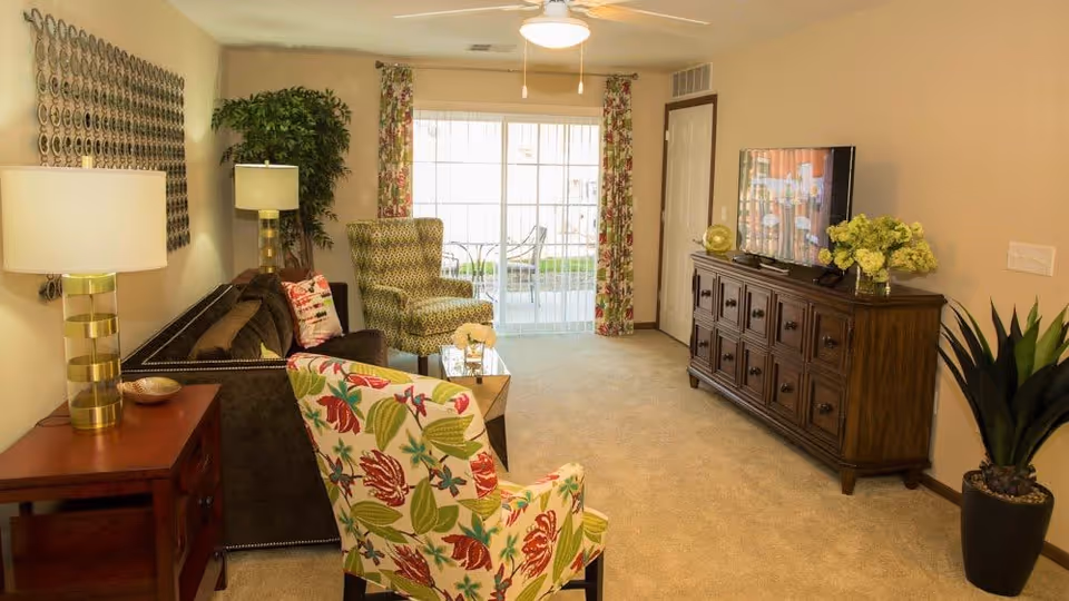 A cozy living room with beige walls and carpet, featuring a dark brown sofa with decorative pillows, a floral patterned armchair, a green patterned armchair, a wooden side table with a lamp, a wooden TV cabinet with a flat screen TV, and a large potted plant. Sliding glass doors with floral curtains lead to an outdoor patio with chairs and a table.