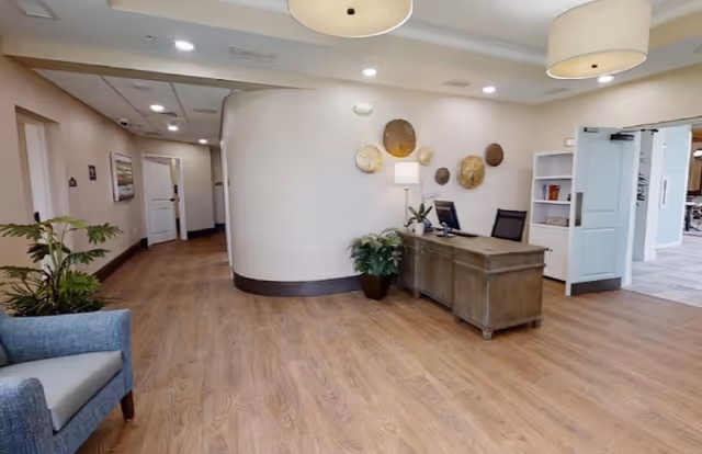 Interior view of a reception area in a senior living facility with a wooden desk, computer, potted plants, decorative wall art, and a hallway leading to other rooms. The space has wood flooring, light-colored walls, and ceiling lights.