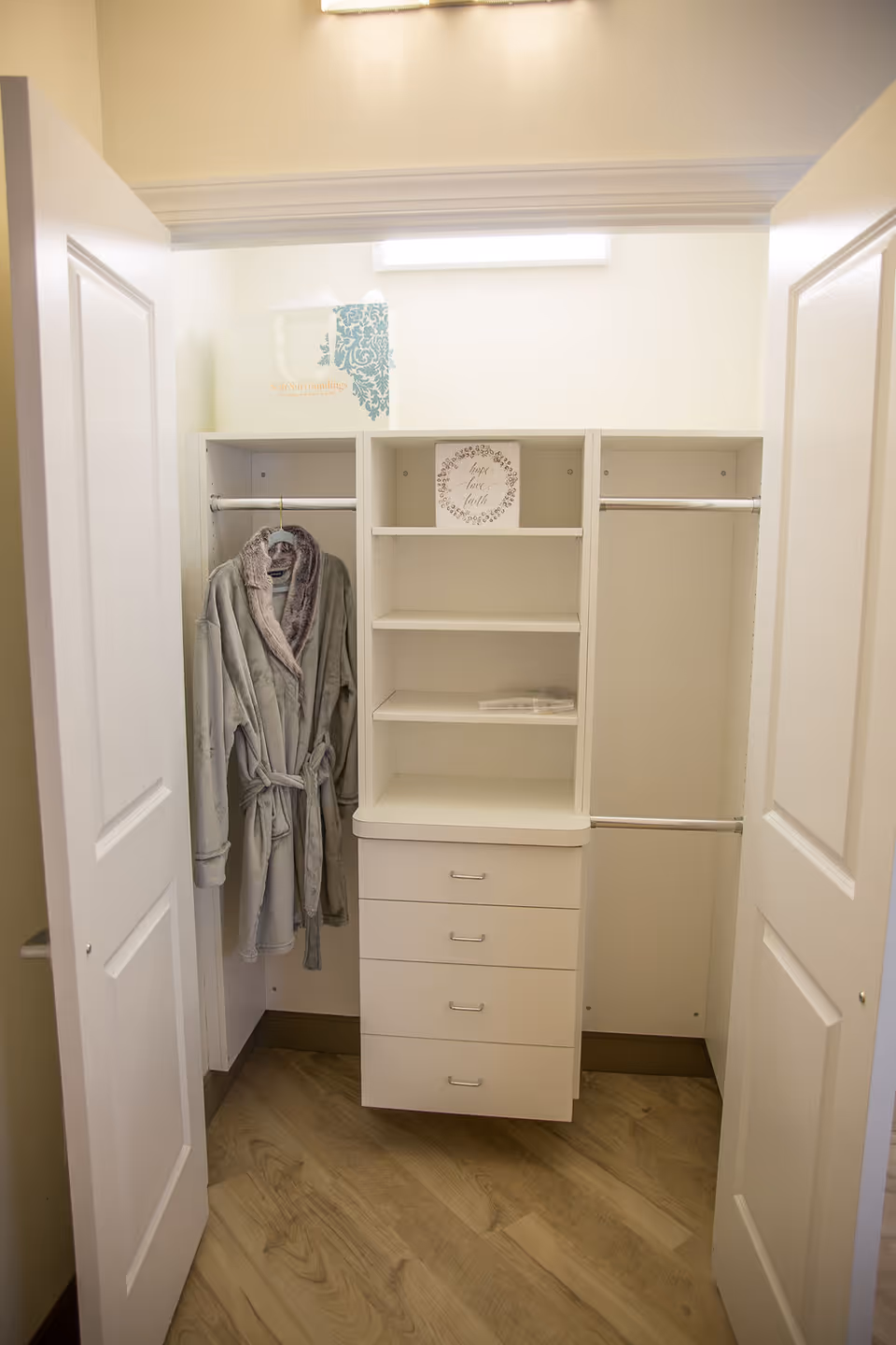 Open closet with white shelves and drawers, a gray bathrobe hanging on the left side, and a decorative sign on the middle shelf. The closet has double doors and wood flooring.