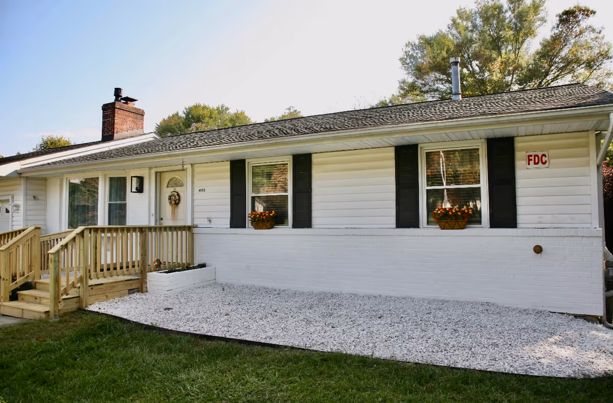 Exterior view of a single-story white house with a shingled roof, two windows with flower boxes, a wooden ramp leading to the front door, and a small garden area covered with white gravel. Trees are visible in the background under a clear sky.