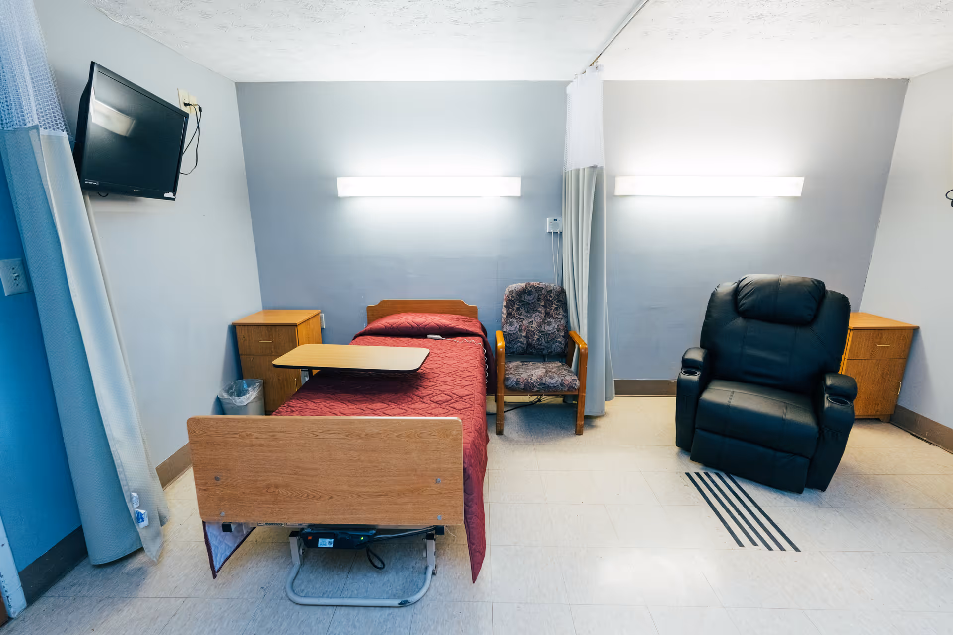 A small senior-care room with a single bed covered in a burgundy blanket, an overbed table, a patterned armchair, a black recliner, wall-mounted TV and bedside cabinets.