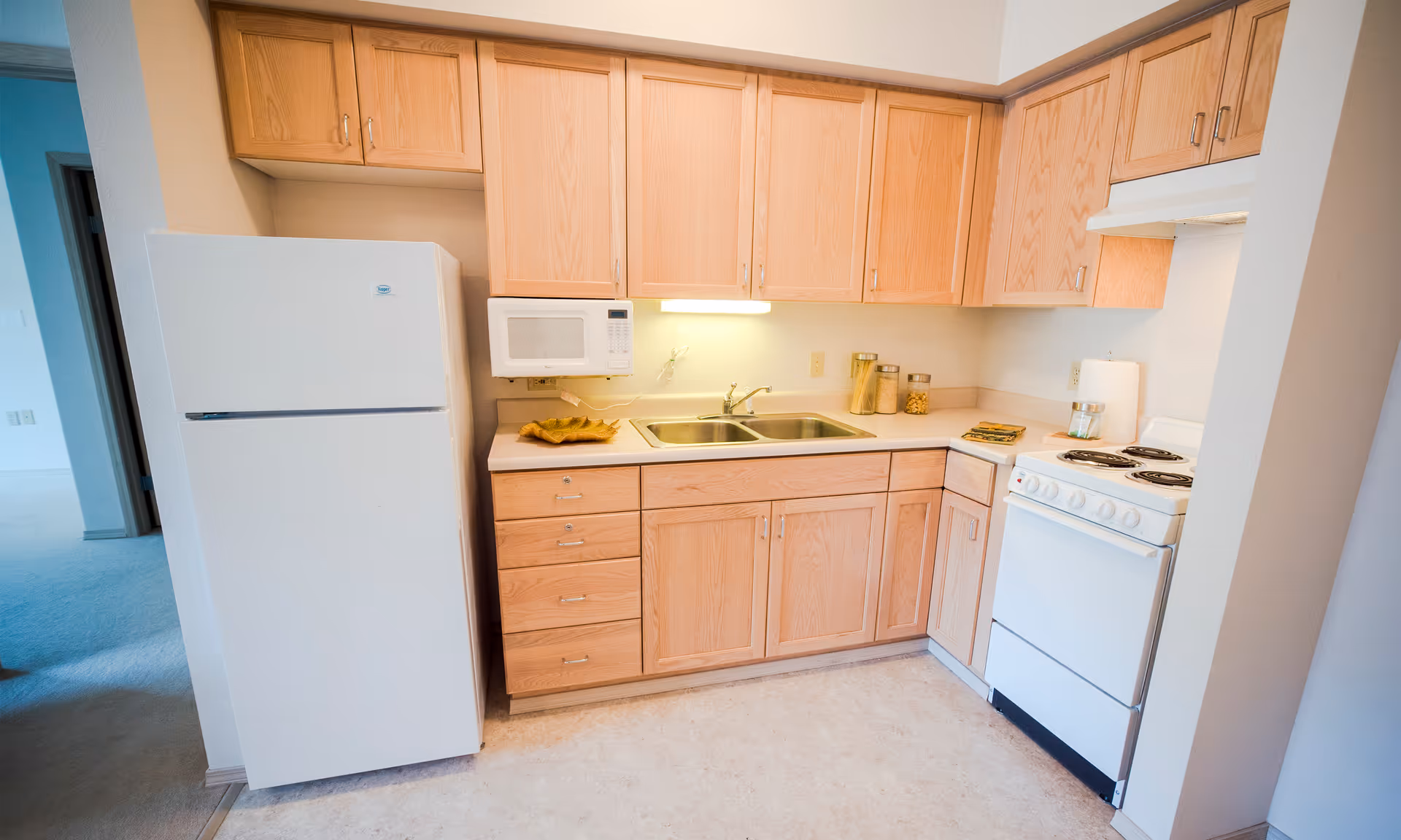 A compact kitchen with light wood cabinets, a white refrigerator, a white microwave mounted above the counter, a double sink, and a white electric stove with four burners. The countertop has a few decorative items including jars and a small dish. The kitchen floor is light-colored and the adjacent room has carpeted flooring.