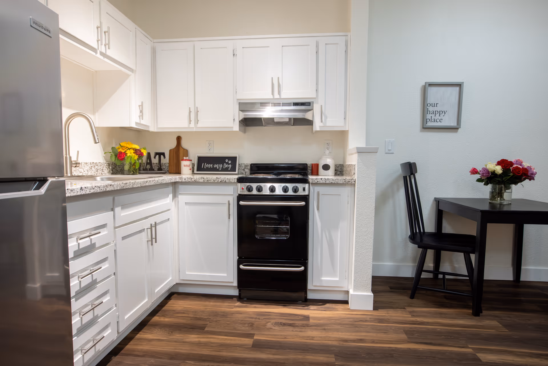 A modern kitchen with white cabinets, a stainless steel refrigerator, a black stove, and granite countertops. There is a vase with colorful flowers on the counter near the sink and a small dining area with a black chair and table holding a vase of flowers. A framed sign on the wall reads 'our happy place.'