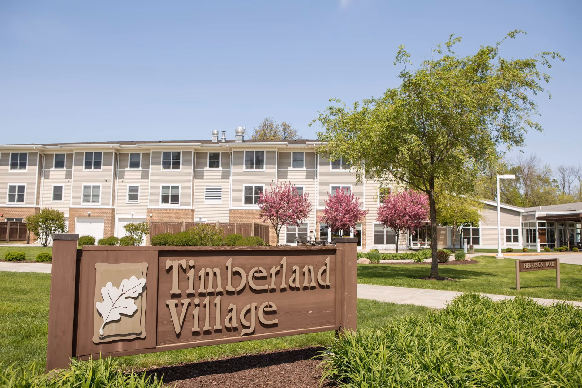 Exterior view of Timberland Village Senior Living facility with a large wooden sign in the foreground displaying the name. The building is a multi-story structure with beige siding and multiple windows. The surrounding area features green grass, trees with pink blossoms, and a clear blue sky.