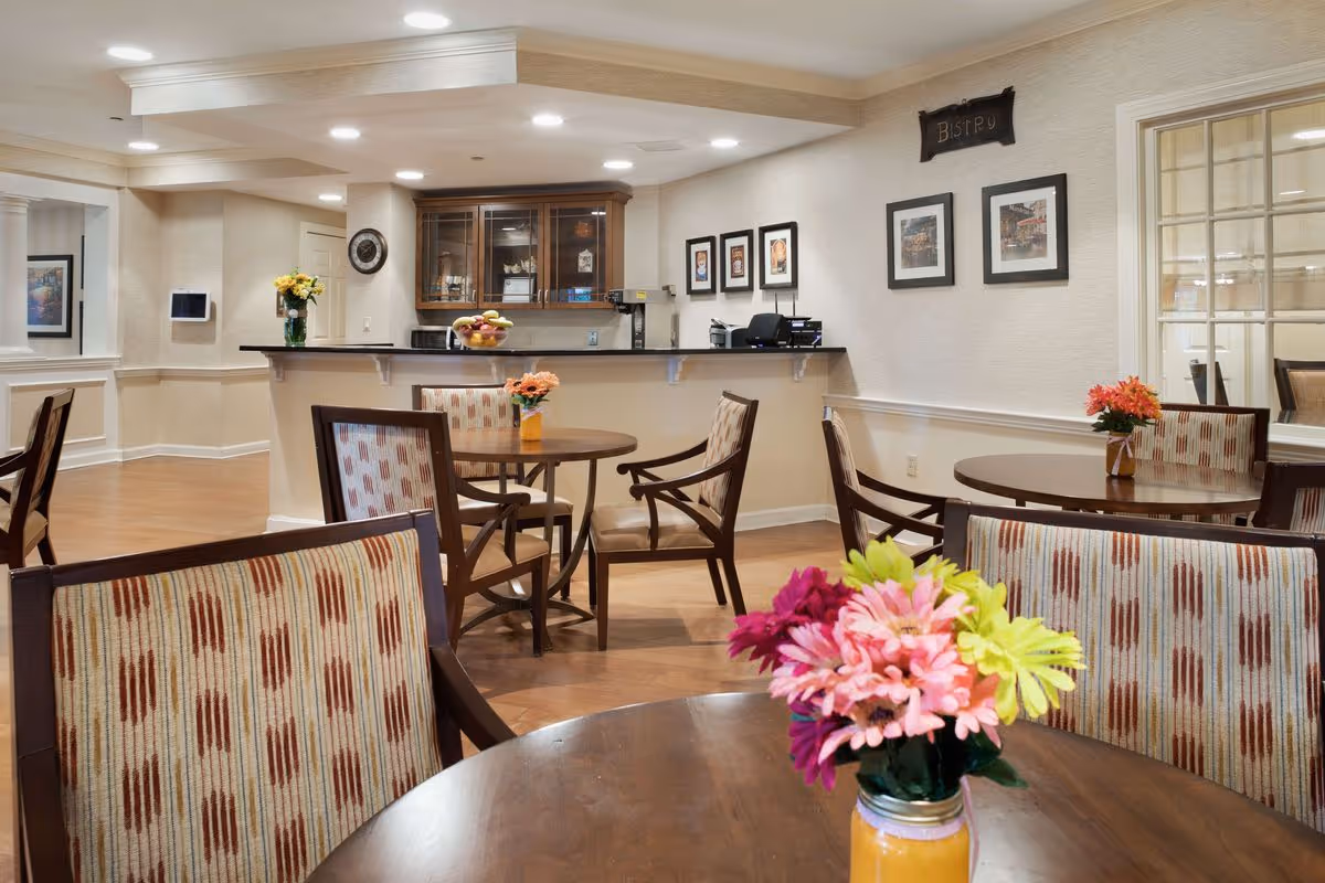 Interior view of a dining area in a senior living facility named Sunrise of Wayland. The room features round wooden tables with patterned cushioned chairs. Each table has a small vase with colorful flowers. In the background, there is a counter with a fruit bowl and a cabinet with glass doors. The walls are decorated with framed pictures and a small sign that reads 'Bistro'. The space is well-lit with recessed ceiling lights and has a warm, inviting atmosphere.