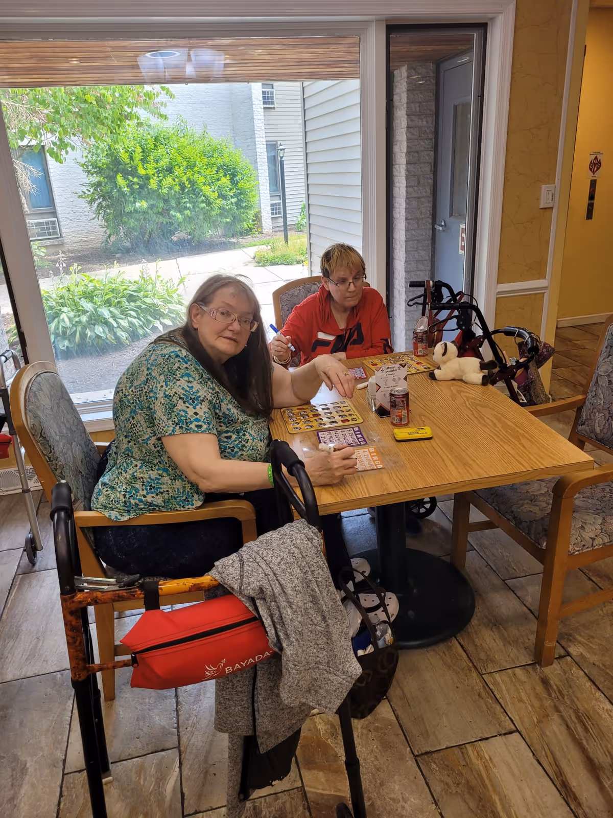 Two elderly women sitting at a wooden table playing bingo in a room with large windows showing greenery outside. One woman is wearing a green patterned shirt and glasses, the other is in a red shirt. There are walkers next to them and bingo cards on the table along with drinks and a small stuffed animal.
