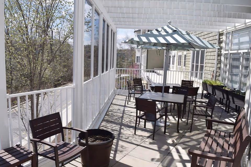 A covered outdoor patio area with several dark wooden chairs and tables, including one table with a large umbrella. The patio is enclosed with white railings and has a view of trees and neighboring buildings. There are also planter boxes with greenery along one side.