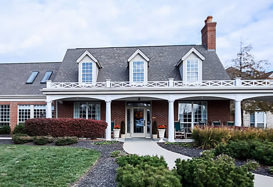 Front exterior view of a brick building with a gray shingled roof, three dormer windows, white columns supporting a porch, and a walkway leading to double glass doors. The landscaping includes green bushes, red shrubs, and a well-maintained lawn.