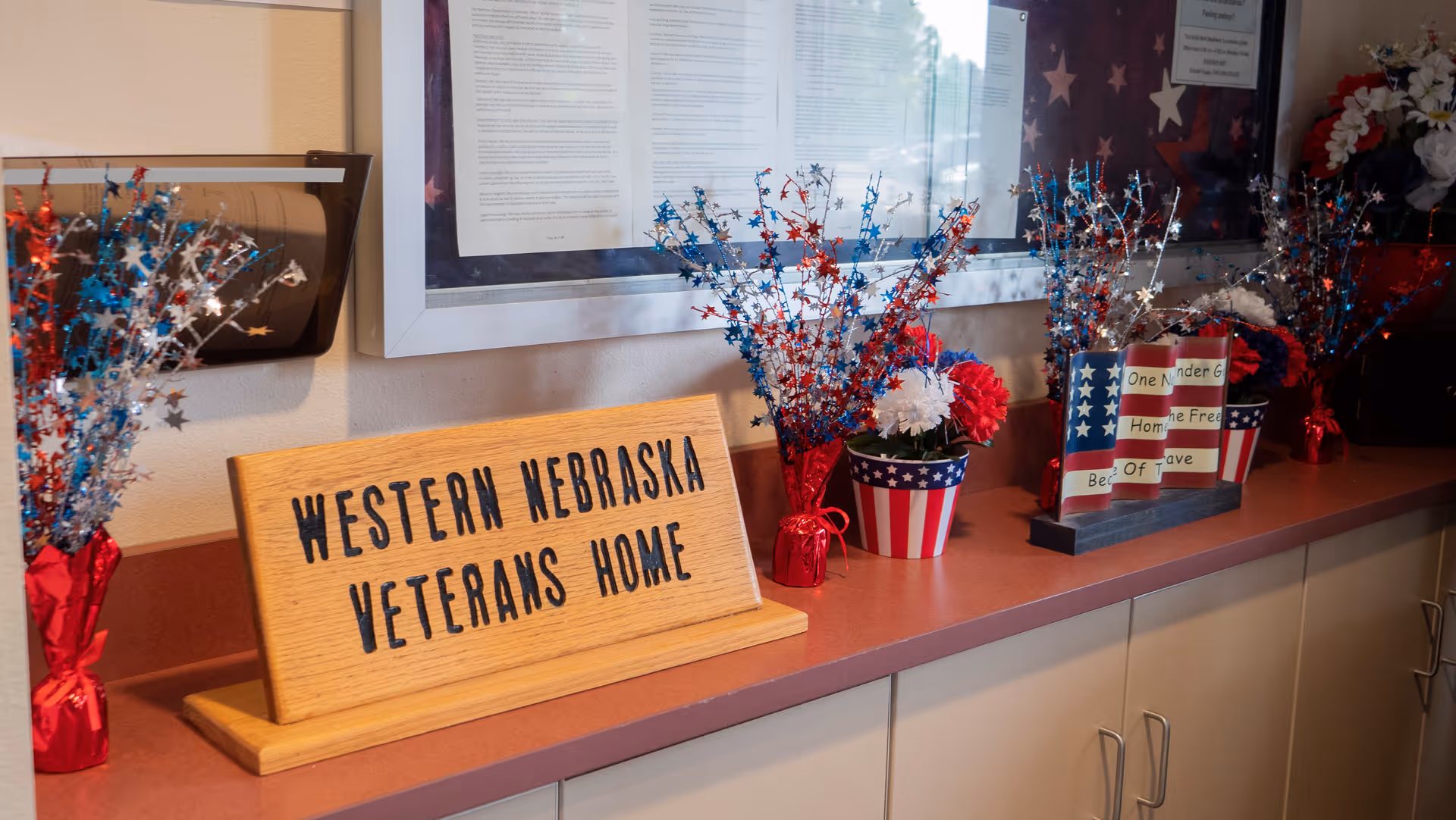 A countertop display at Western Nebraska Veterans Home featuring a wooden sign with the facility name, patriotic red, white, and blue star decorations, and a small American flag-themed flower pot. Behind the display is a framed document and part of an American flag.
