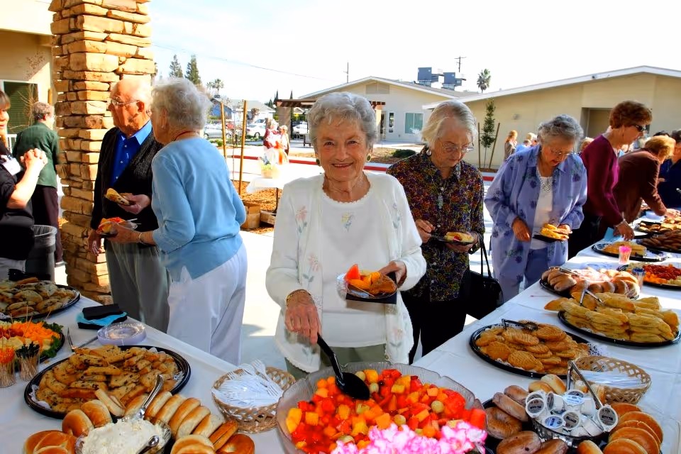 A group of elderly people serving themselves food from a buffet table set up outdoors at Sierra View Homes Retirement Community. The table is filled with various pastries, cookies, bread rolls, and a large bowl of mixed fruit. The setting appears sunny with buildings and trees in the background.