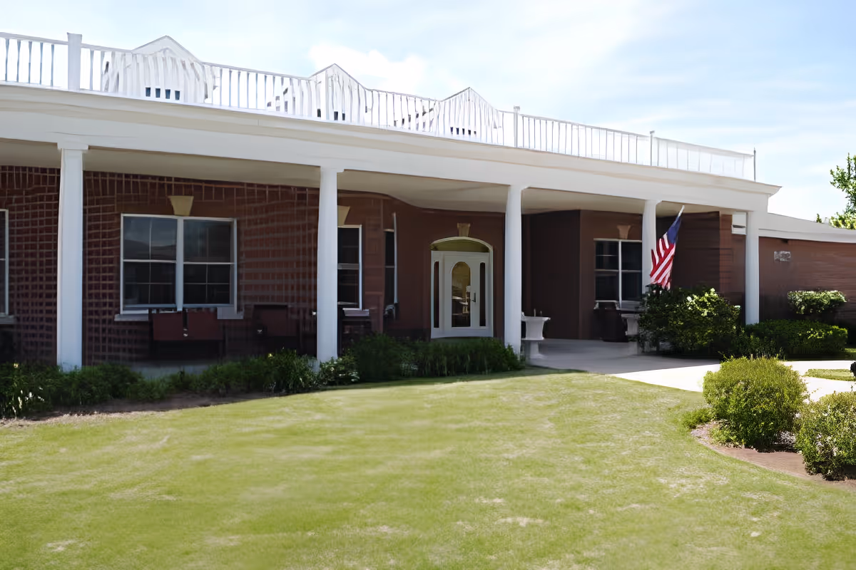 Exterior view of a single-story brick building with white columns supporting a covered porch. There are several windows and a white door with glass panels. An American flag is displayed near the entrance, and there are bushes and a well-maintained lawn in front of the building under a partly cloudy sky.