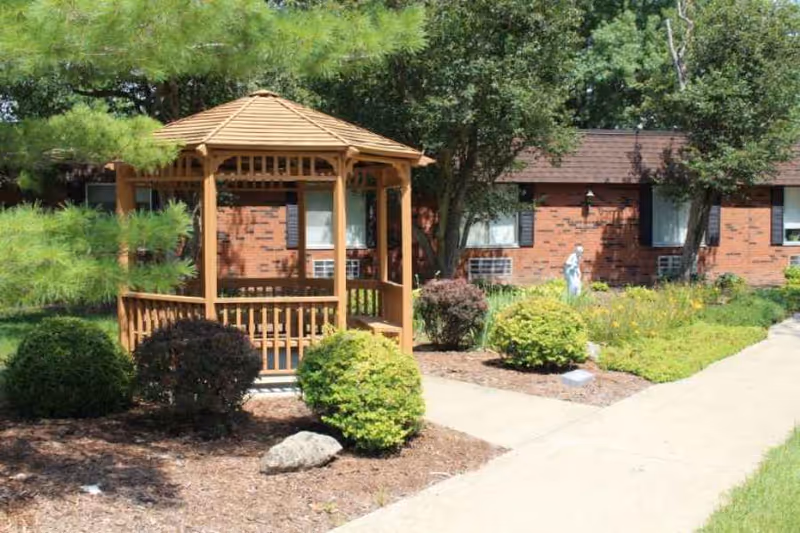 A wooden gazebo surrounded by bushes and trees in a garden area with a paved walkway leading to a brick building with windows and air conditioning units.