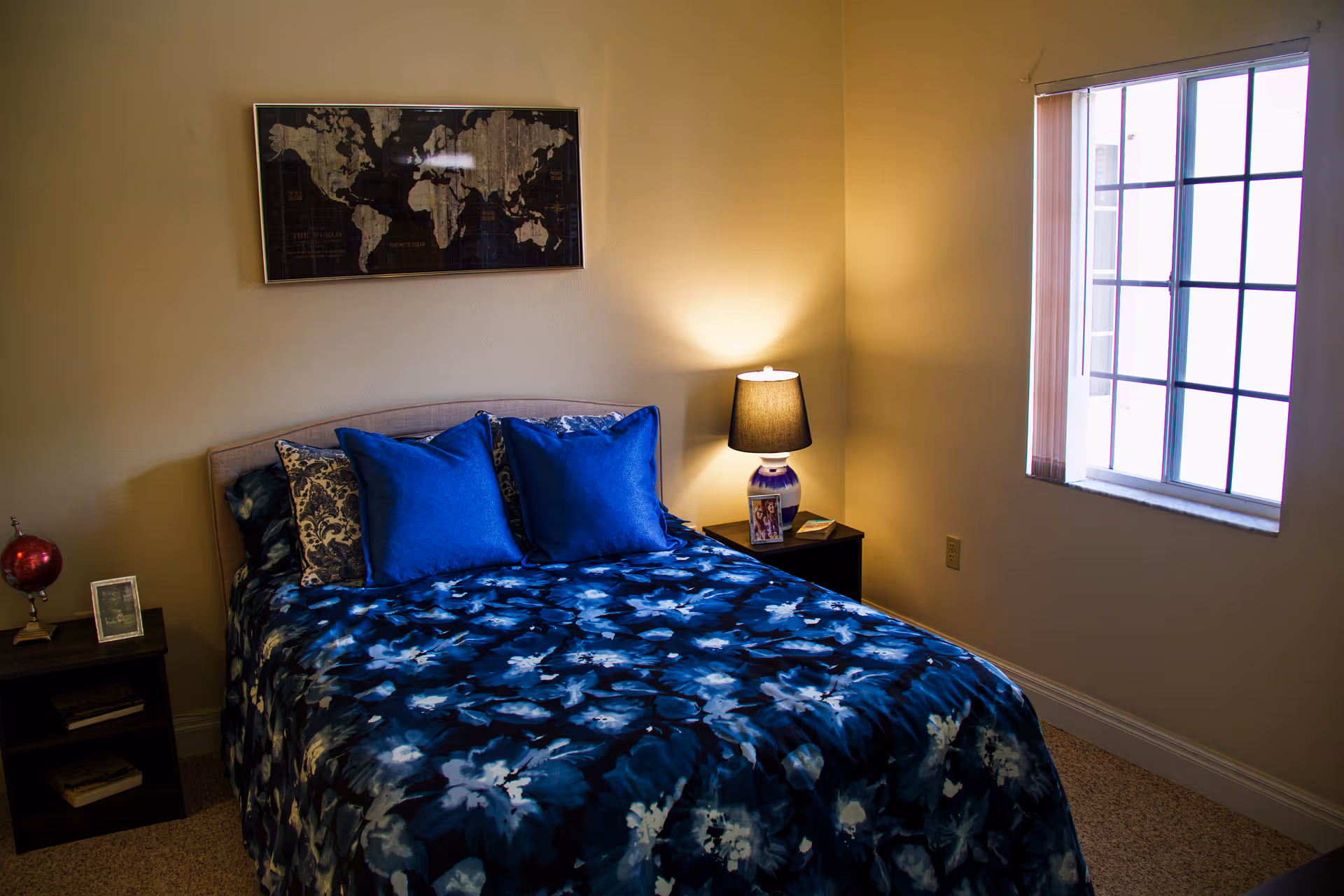 A cozy bedroom with a bed covered in a blue floral bedspread and matching blue pillows. There is a nightstand on each side of the bed, one with a lamp and a framed photo, and the other with a small globe and a framed picture. A window with vertical blinds allows natural light into the room, and a framed world map hangs on the wall above the bed.