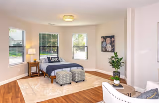 A bright and spacious bedroom with a bed featuring a dark blue comforter and a pillow with the word 'love'. The room has three windows with white blinds, a wooden nightstand with a lamp, two patterned ottomans at the foot of the bed, a large beige area rug, a round wooden coffee table with a plant, a white armchair, and a floral painting on the wall.