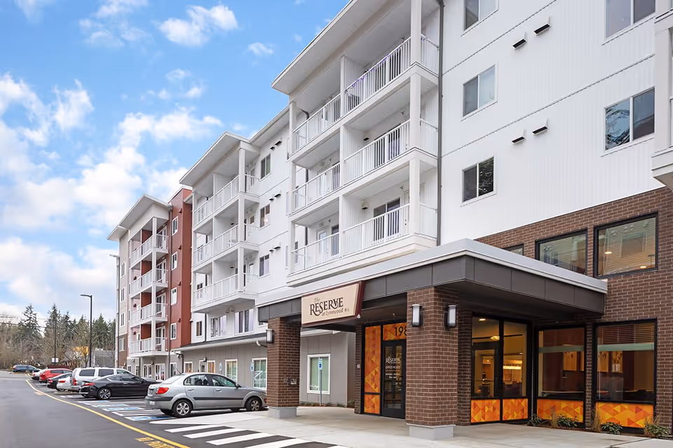 Exterior view of The Reserve At Lynnwood senior living facility showing a multi-story building with balconies, a brick and white facade, a parking area with several cars, and a clear blue sky with some clouds.