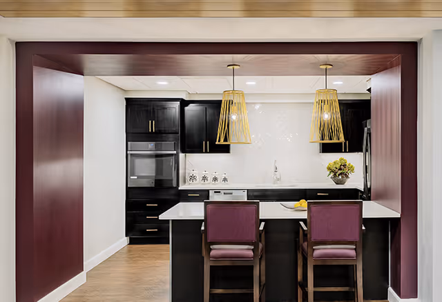 Modern kitchen with dark cabinetry, a white countertop island with two purple cushioned chairs, two pendant lights with gold wireframe shades hanging above the island, and a white tiled backsplash. The kitchen features built-in appliances and a decorative plant on the counter.