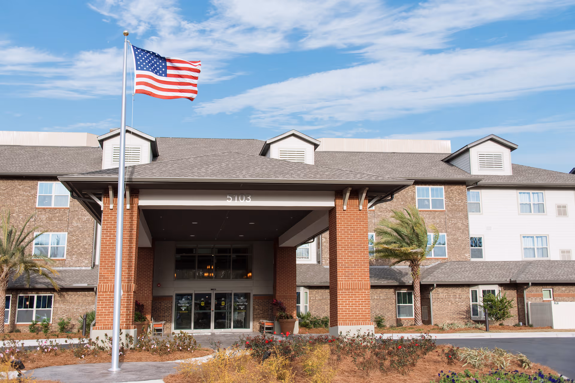 Front entrance of a multi-story senior living building with an American flag on a pole and landscaped grounds.