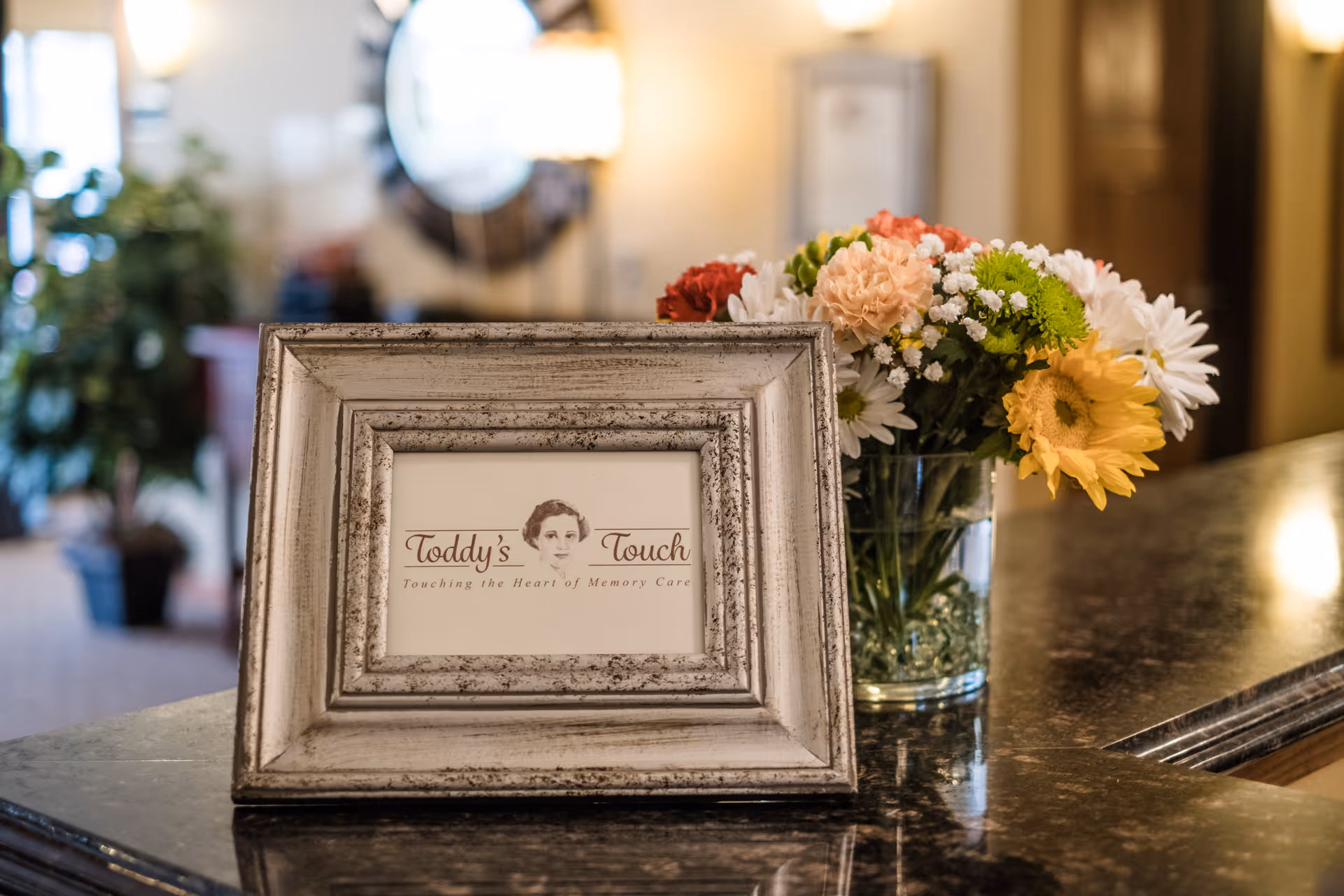 A framed sign on a countertop with the text 'Toddy's Touch Touching the Heart of Memory Care' next to a vase of colorful flowers including daisies and carnations, with a softly lit interior background.