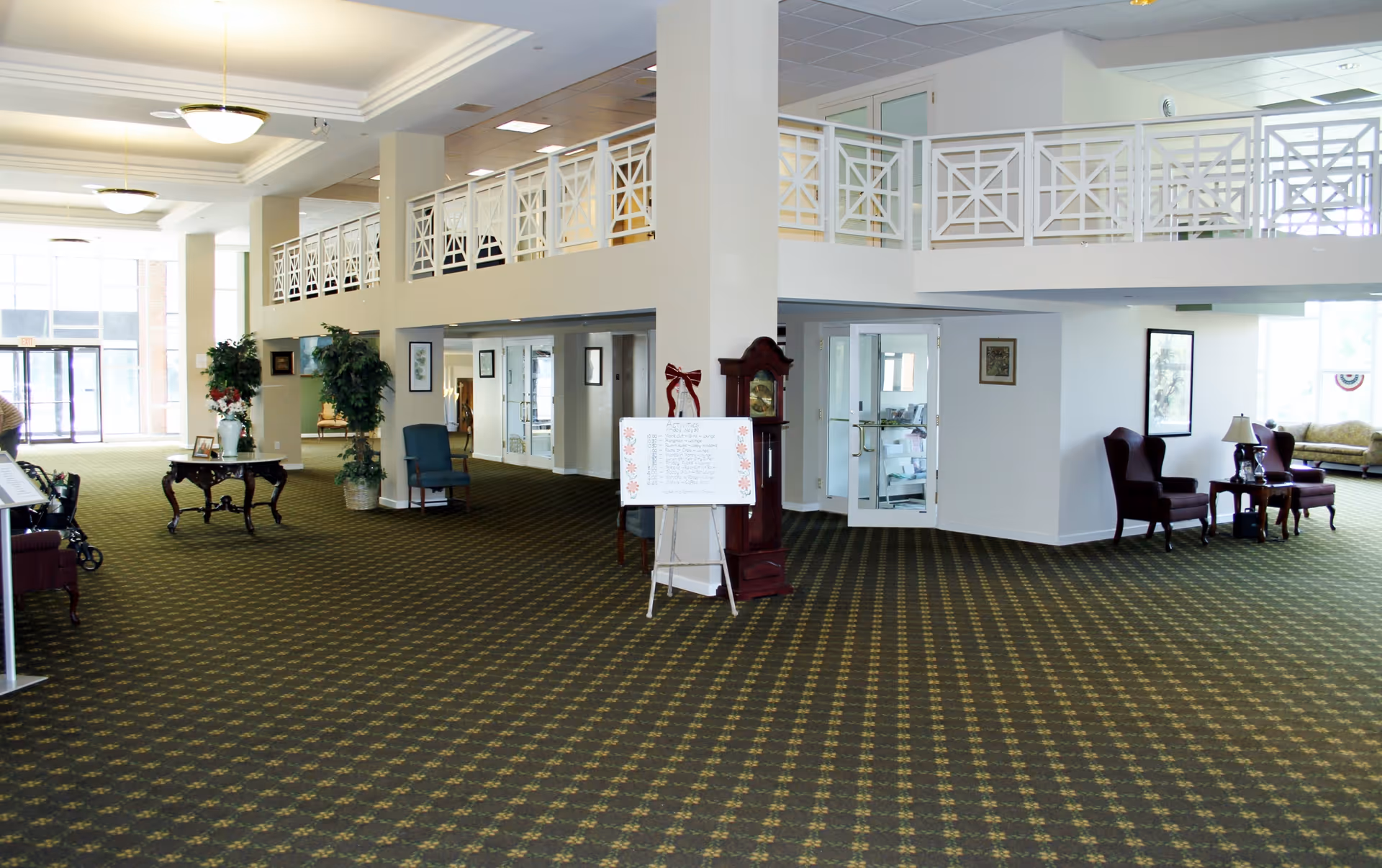 Spacious interior common area of a senior living facility with patterned carpet, white walls, and a high ceiling. The area features a grandfather clock, a whiteboard on an easel, several chairs, small tables with lamps, potted plants, and a balcony with white railings overlooking the space. Large windows allow natural light to fill the room.