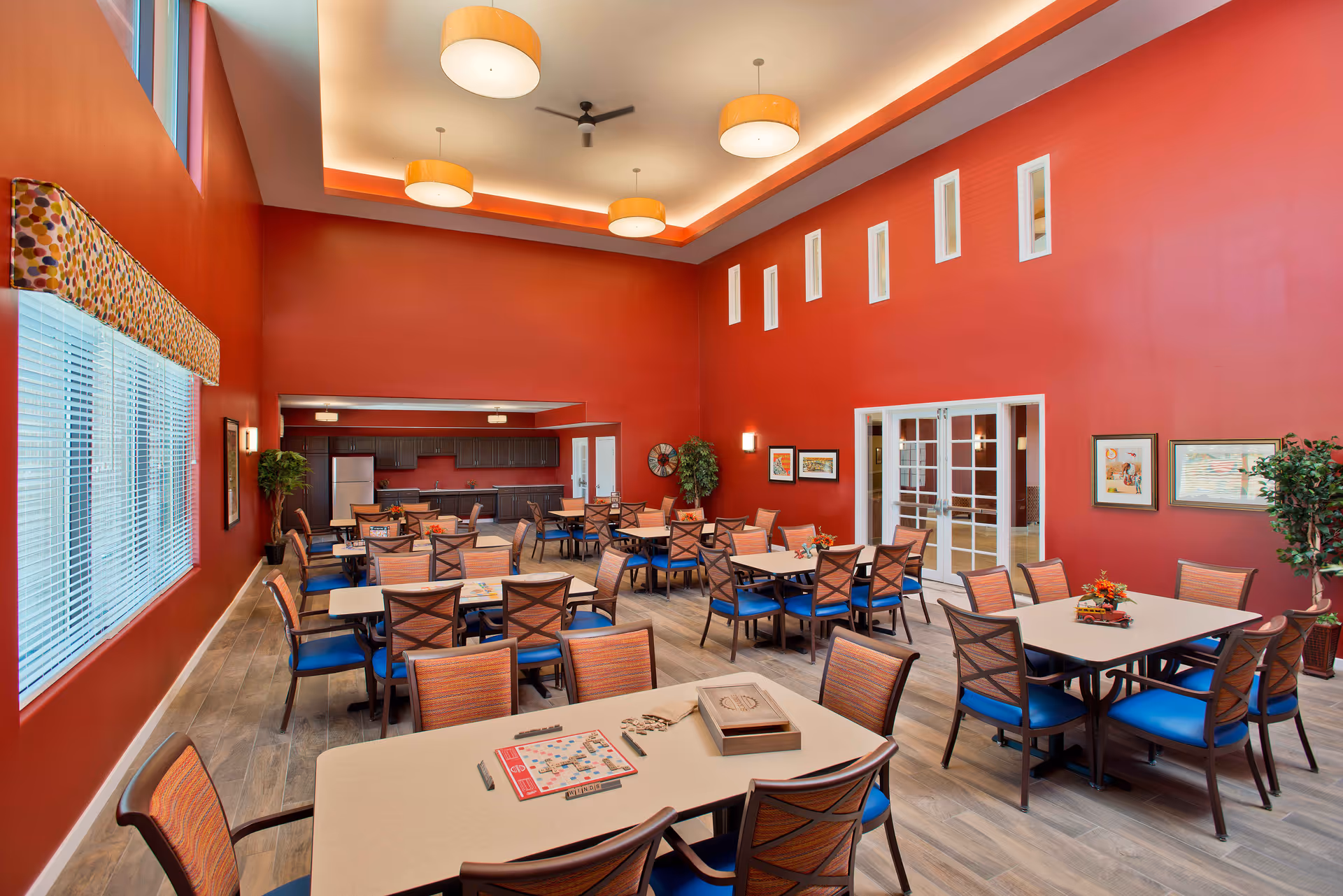 A spacious community room with red walls and a high ceiling featuring four round ceiling lights and a ceiling fan. The room contains multiple tables with chairs arranged around them, some tables have small decorative items and a Scrabble game set up. Large windows with blinds are on one side, and there are framed pictures and plants decorating the room. In the background, there is a kitchenette area with dark cabinets and a refrigerator.