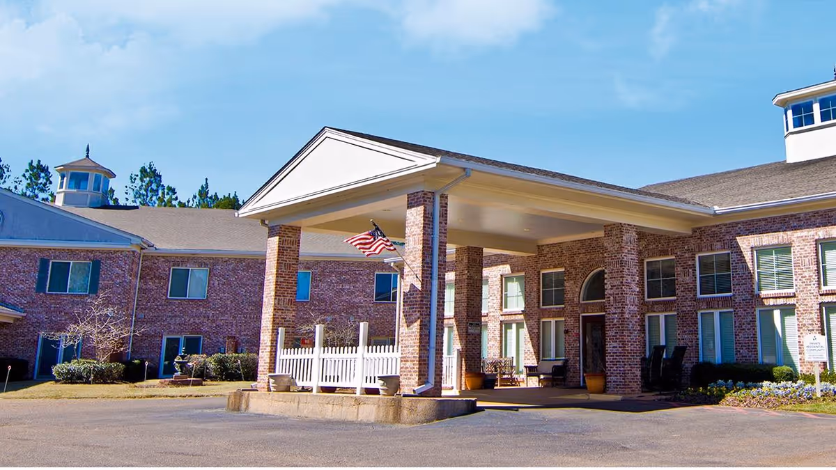Front entrance of a two-story brick senior living facility with a covered portico, American flag, and outdoor seating.