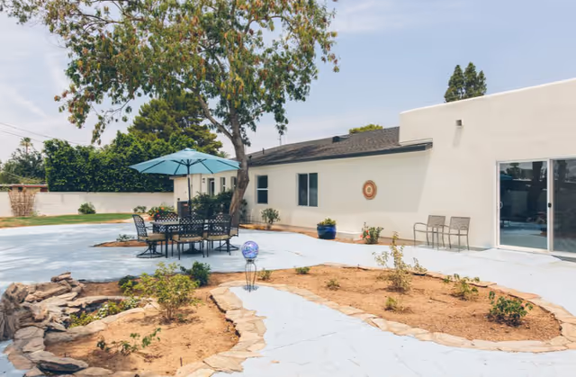 Outdoor patio area with a round table and six chairs under a large blue umbrella. The patio is surrounded by a garden bed with small plants and a tree. The building in the background is white with several windows and a sliding glass door.