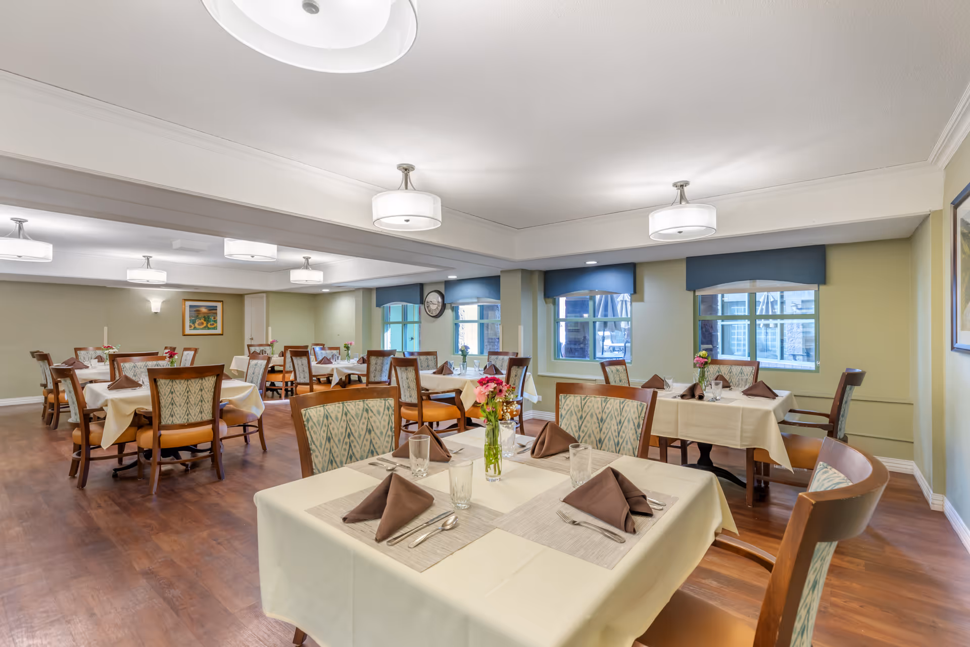 A dining room in a senior living facility with multiple tables covered in cream-colored tablecloths, each set with brown folded napkins, glasses, and silverware. The chairs have wooden frames with patterned upholstery. There are windows with blue valances along one wall, and the room is lit by ceiling lights. A vase with flowers is placed on the table in the foreground.