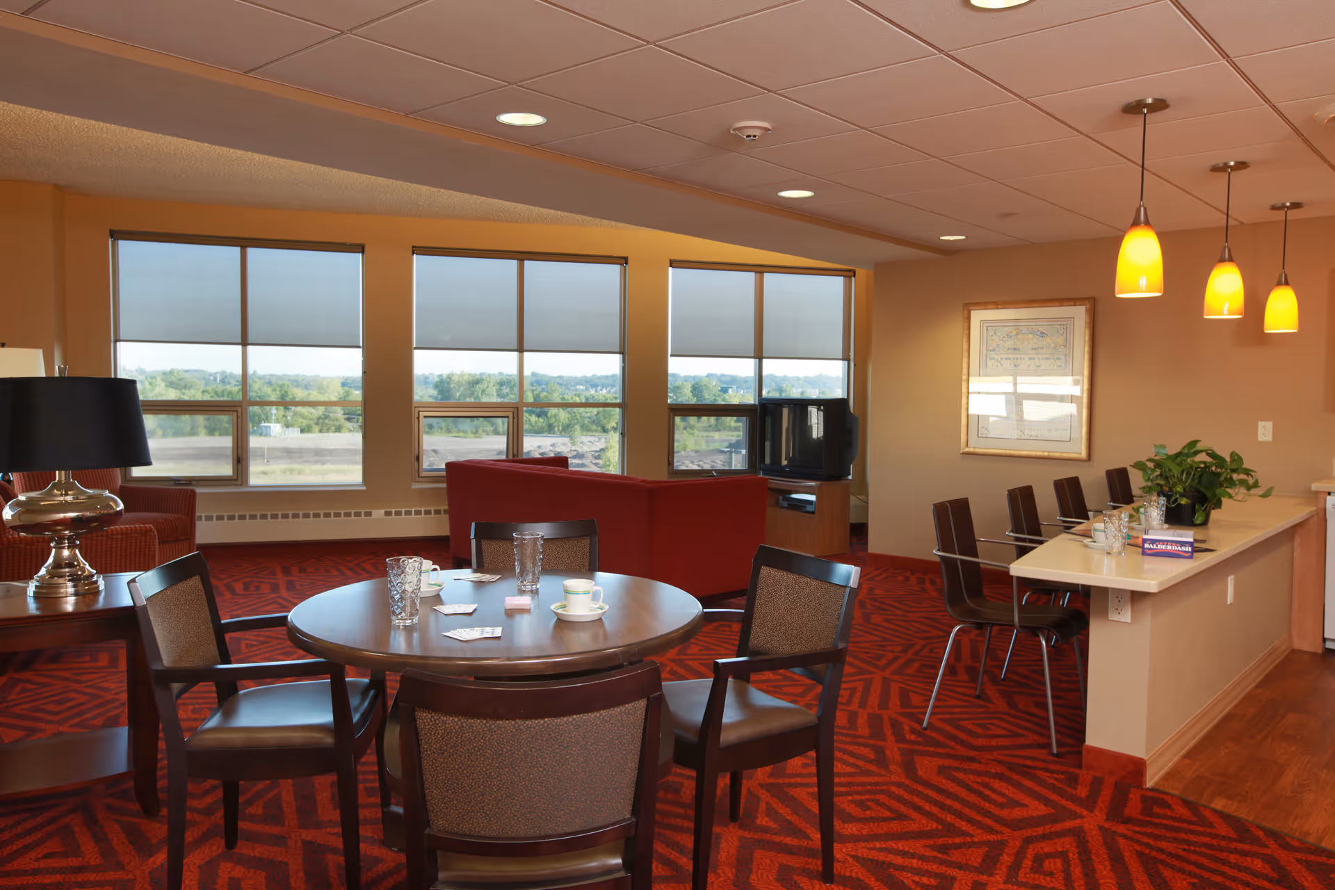 A spacious living room area with large windows showing an outdoor view. The room features a round table with four chairs, a red couch, a TV on a stand, a lamp on a side table, and a counter with bar stools and hanging pendant lights. The floor is covered with a red patterned carpet.