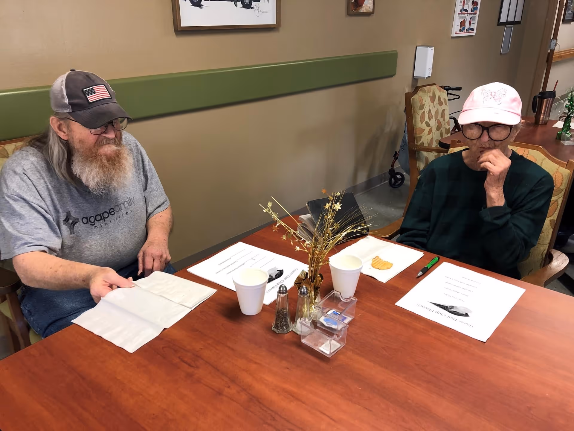 Two elderly people seated at a table with papers, cups, and a small centerpiece in a communal dining/activity room.
