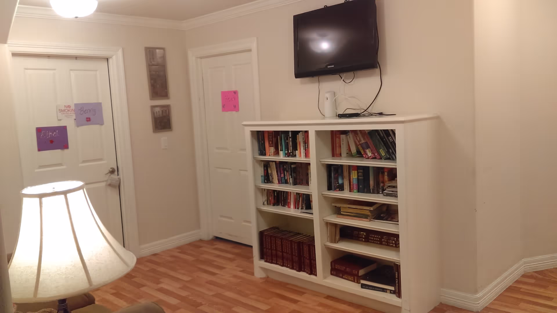 Interior view of a room with light-colored walls and wooden flooring. There is a white bookshelf filled with books against the wall, and a flat-screen TV mounted above it. Two closed white doors are visible, each with colorful paper signs attached. A lit table lamp with a white lampshade is partially visible in the foreground.