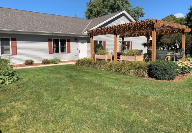 Single-story light gray building with a green lawn and a wooden pergola seating area in front.