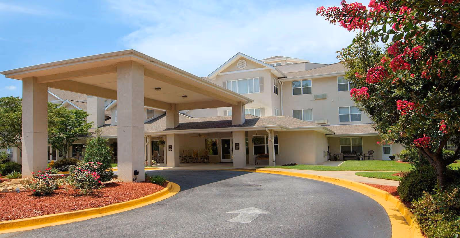 Front entrance of a multi-story senior living facility with a covered porte-cochère, landscaped beds, and a circular driveway.