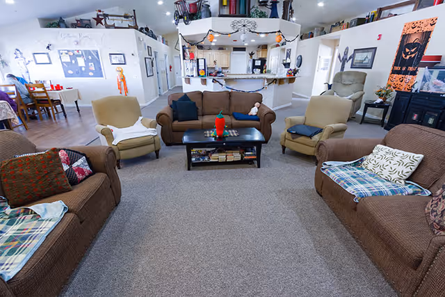 Community living room in a memory care facility with sofas and armchairs arranged around a central coffee table and a kitchenette/reception area in the background.