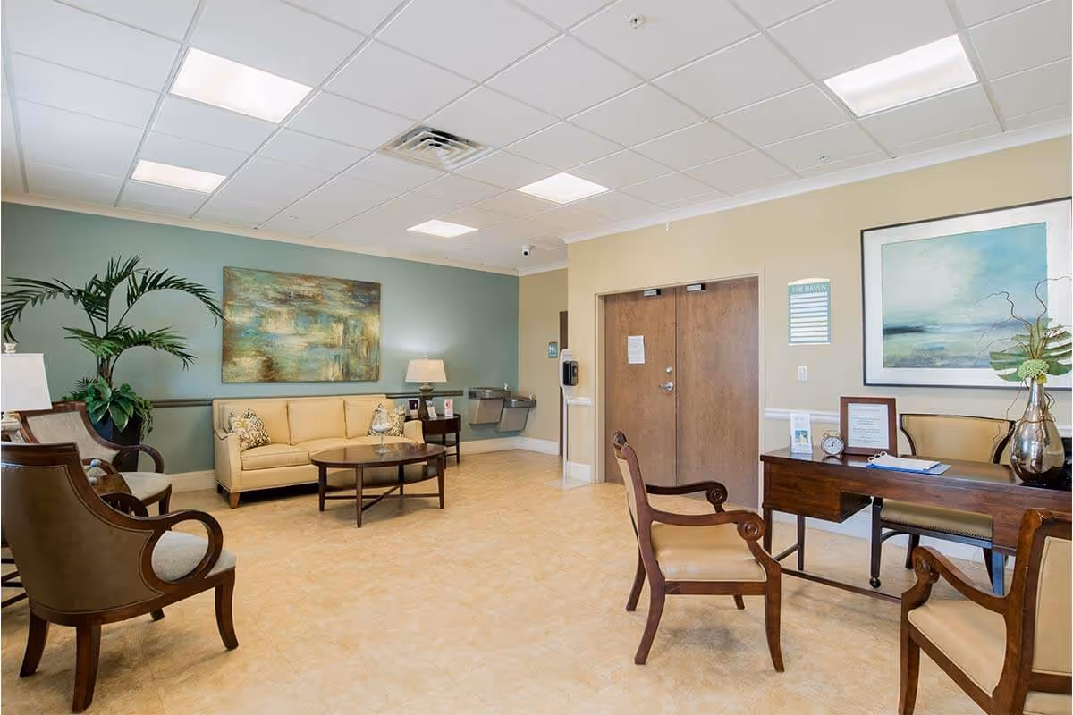 A well-lit waiting or common area with beige tiled flooring, a beige sofa, wooden chairs with beige cushions, a wooden coffee table, and a wooden desk with chairs. The walls are painted in light green and beige tones, decorated with large abstract paintings. There is a water fountain and double wooden doors in the background, along with a potted plant and table lamps providing additional lighting.