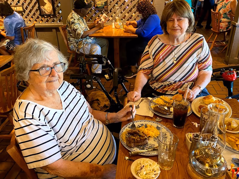Two elderly women sitting at a wooden dining table in a restaurant, enjoying a meal with plates of food including scrambled eggs, meat, and coleslaw. One woman wears glasses and a white shirt with black stripes, while the other wears a striped multicolored top. Other diners are visible in the background.