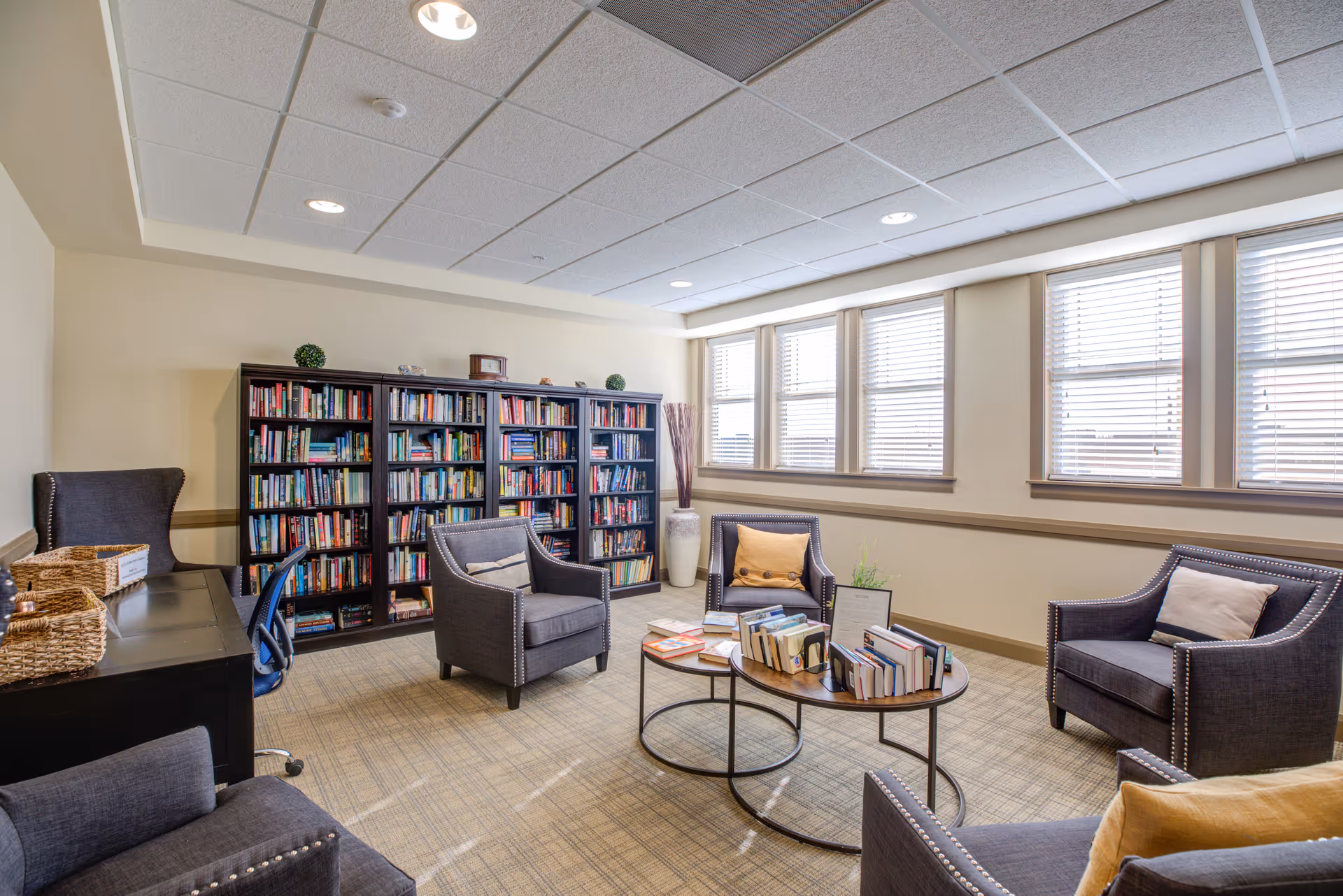 A bright and cozy reading room with four gray armchairs arranged around two nested round wooden tables filled with books. Behind the chairs is a large dark wood bookshelf filled with books and decorative items. The room has beige walls, a carpeted floor, and several windows with white blinds allowing natural light to enter.