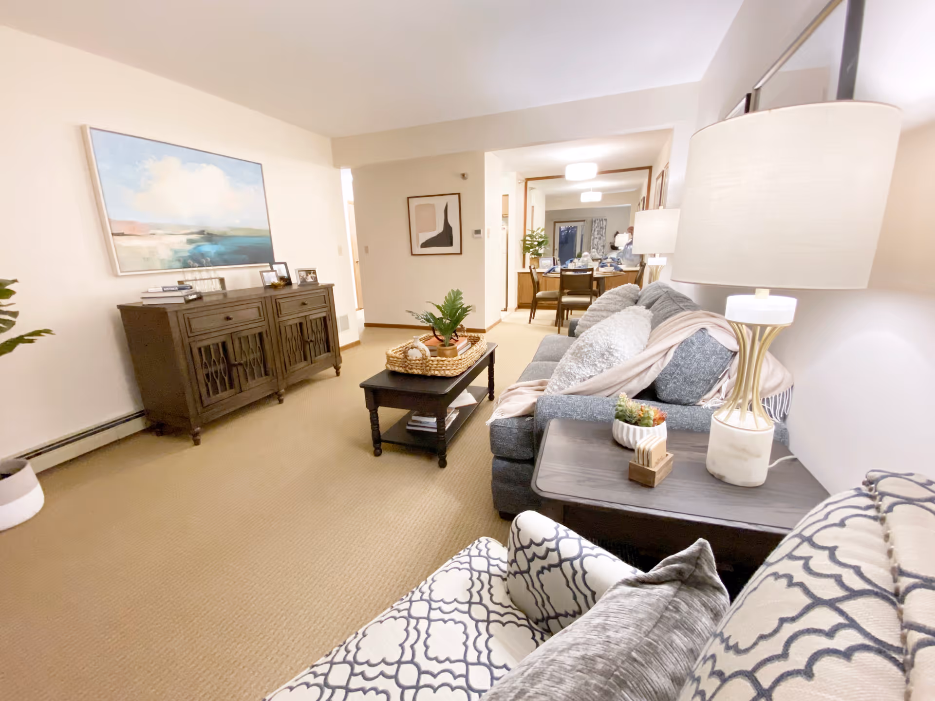 A cozy living room with a gray sofa adorned with pillows and a throw blanket, a patterned armchair, a dark wooden coffee table with a plant centerpiece, and a matching side table with a lamp and small decor items. On the left wall, there is a wooden cabinet with framed photos and a large landscape painting above it. The room opens up to a dining area in the background with a table and chairs, illuminated by ceiling lights.