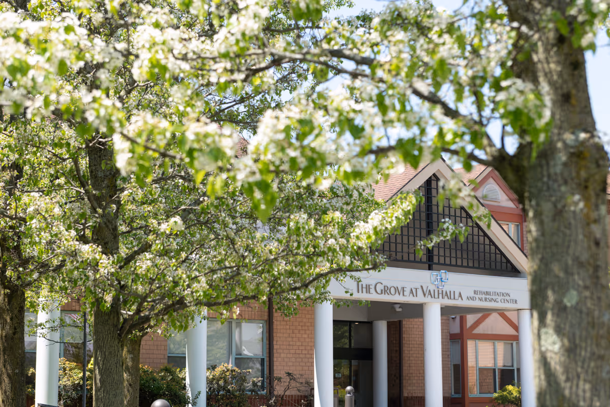 Front entrance of The Grove at Valhalla Rehabilitation and Nursing Center partially obscured by flowering trees.
