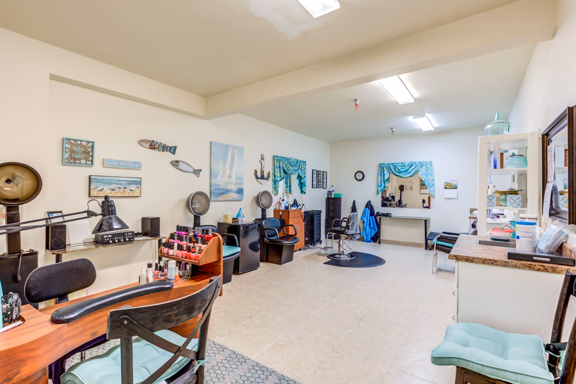 Interior view of a salon area in a senior living facility with multiple styling chairs, a manicure table with nail polish bottles, hair dryers, and decorative wall art including fish and coastal-themed paintings. The room has tiled floors, bright overhead lighting, and a countertop with various supplies.