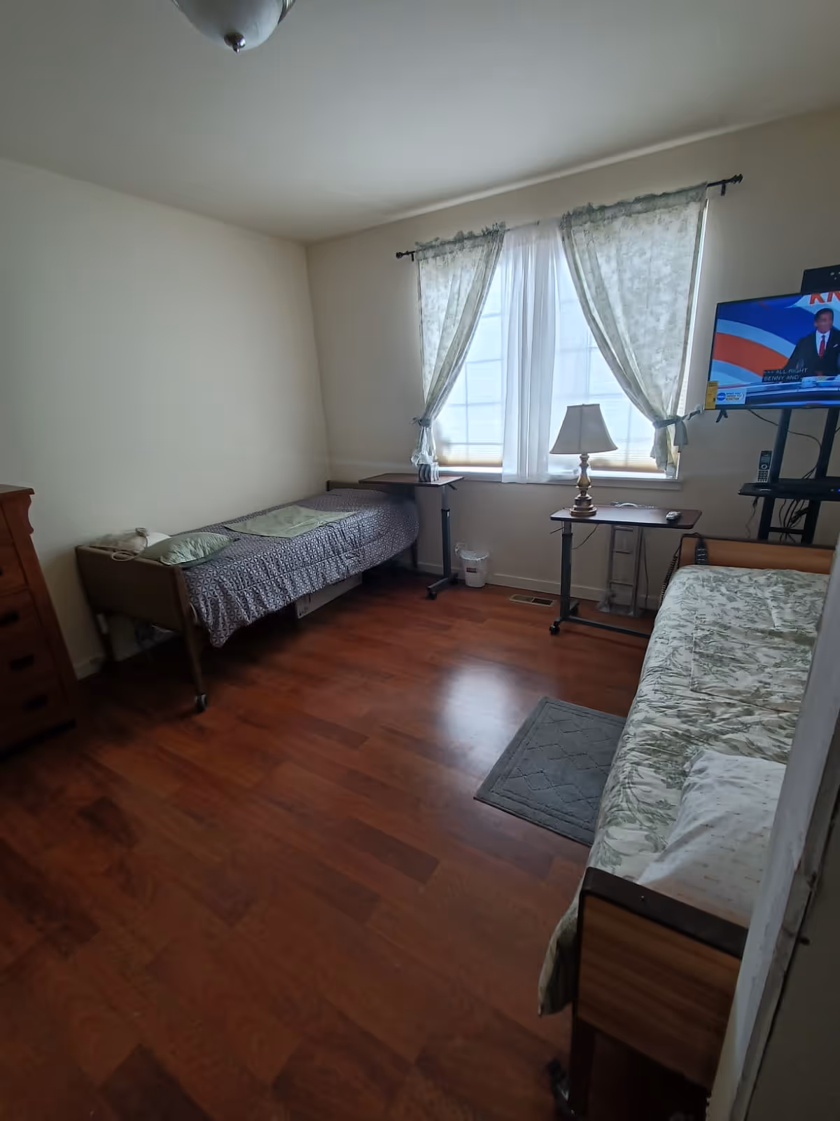 Small shared bedroom with two single beds, a window with light curtains, a lamp and a TV on a stand over wood floors.
