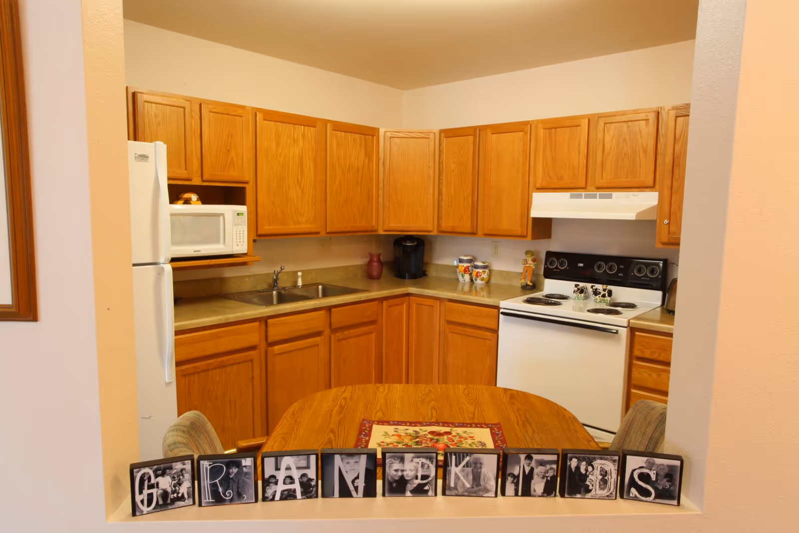 Small kitchen with oak cabinets, white appliances, a sink and stove, and a wooden table with framed photos spelling 'GRANDKIDS' on the pass-through ledge.