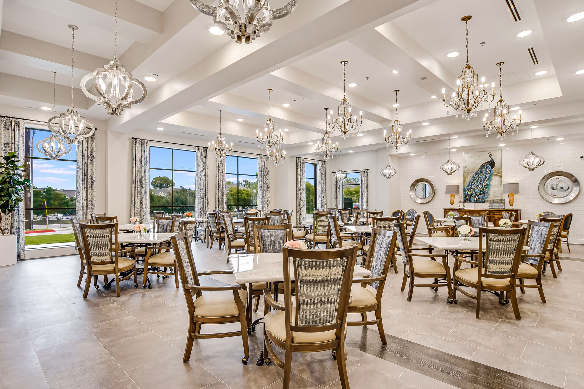 Spacious, elegantly furnished dining room with round tables, wooden chairs, and multiple chandeliers beside large windows.