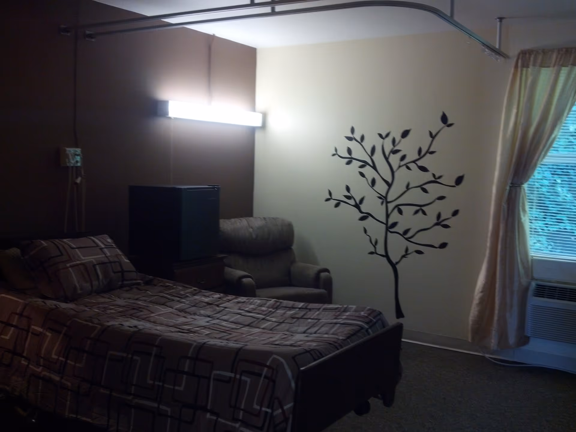 A senior living facility bedroom with a single bed covered in a patterned brown and beige bedspread, a cushioned armchair, a small black refrigerator on a wooden stand, a wall decal of a tree with leaves, a window with beige curtains, and an air conditioning unit below the window.