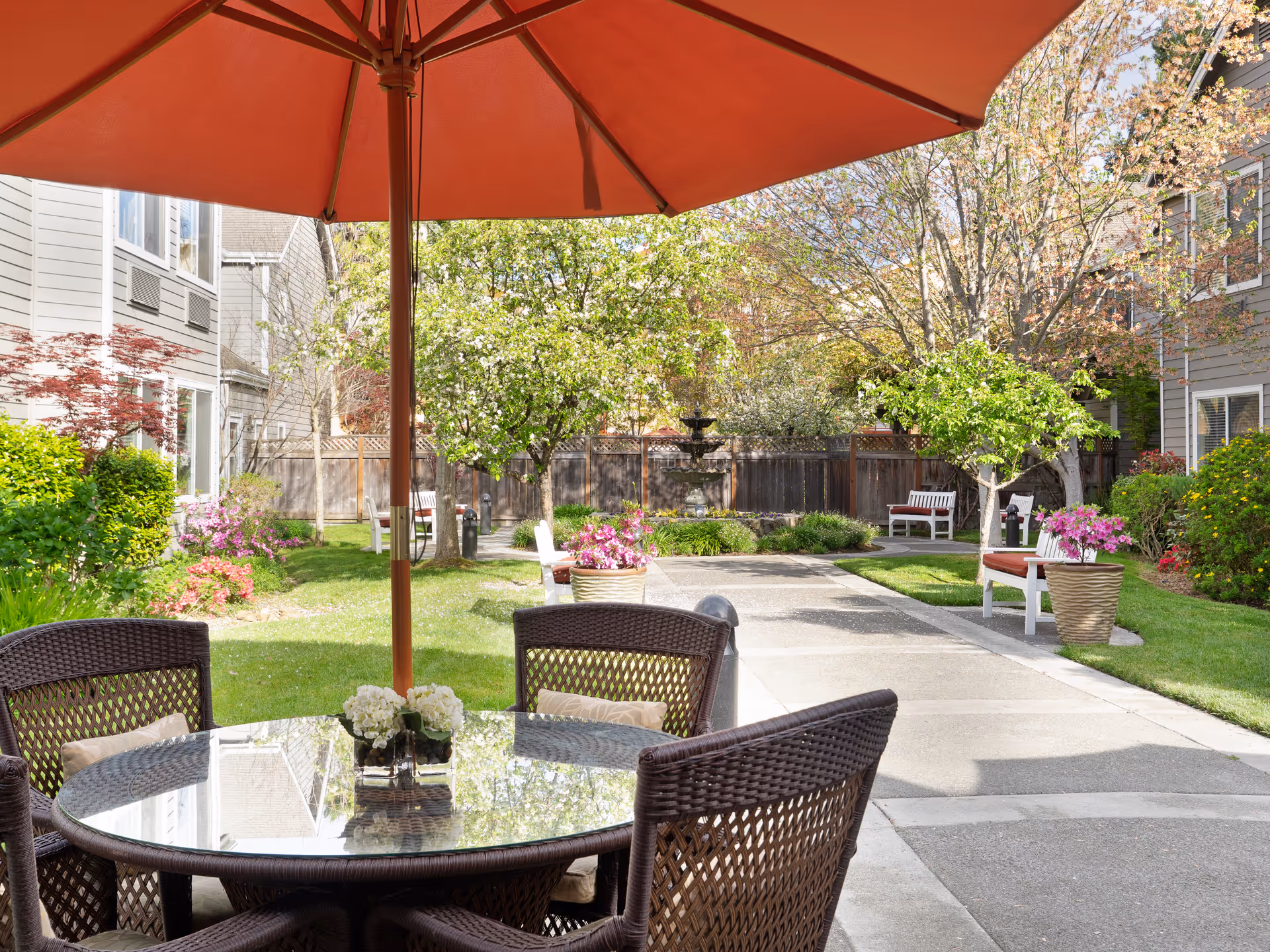 Outdoor patio area at Windsong of Sonoma Senior Living featuring a round glass-top table with four wicker chairs under a large red umbrella. The patio overlooks a garden with green grass, flowering plants, trees, benches, and a water fountain in the background.