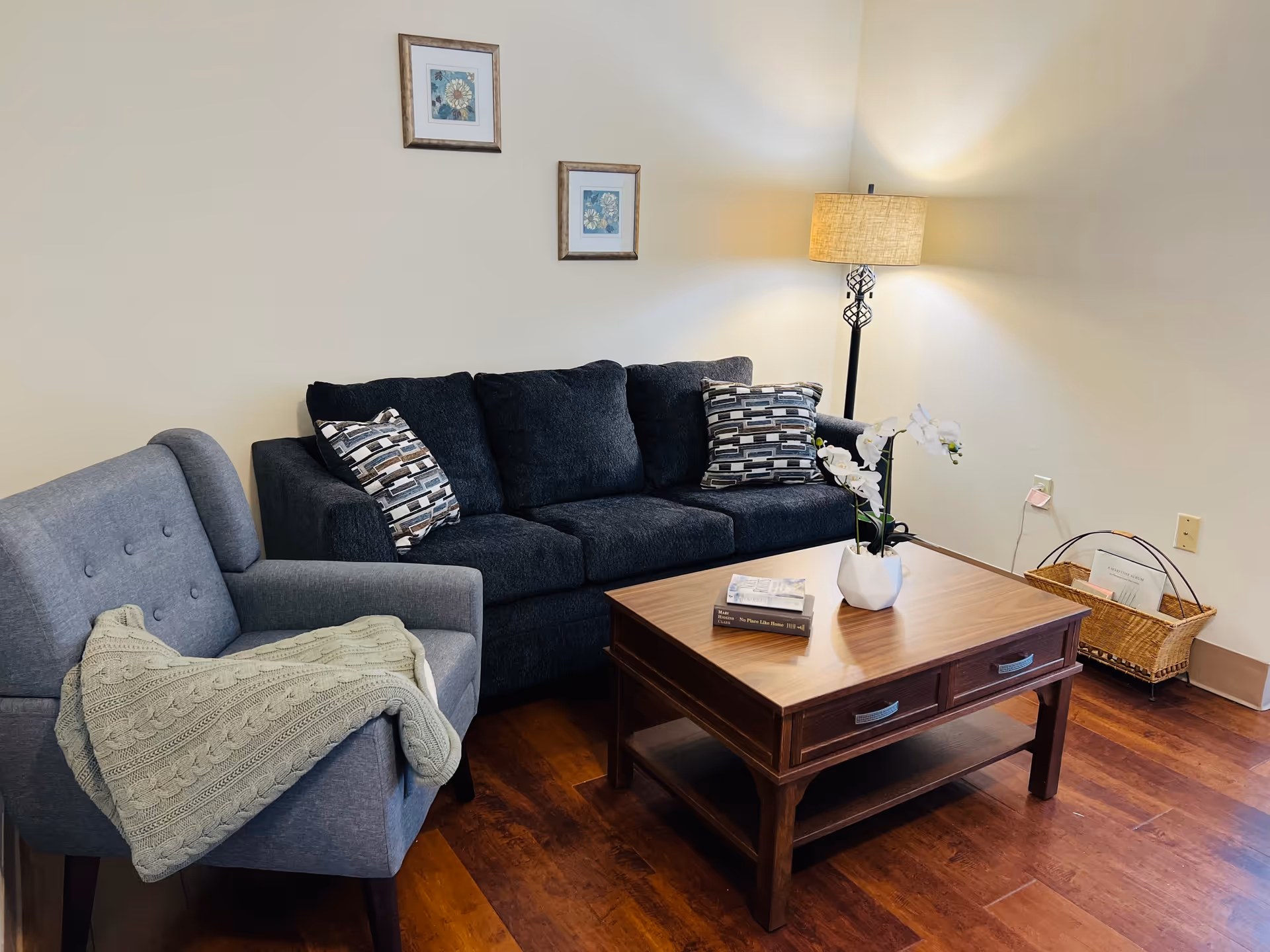 A cozy living room with a dark gray sofa adorned with patterned cushions, a light gray armchair with a knitted throw draped over it, a wooden coffee table with two books and a white orchid plant, a floor lamp with a beige shade, two framed floral artworks on the wall, and a wicker basket holding magazines on the floor.